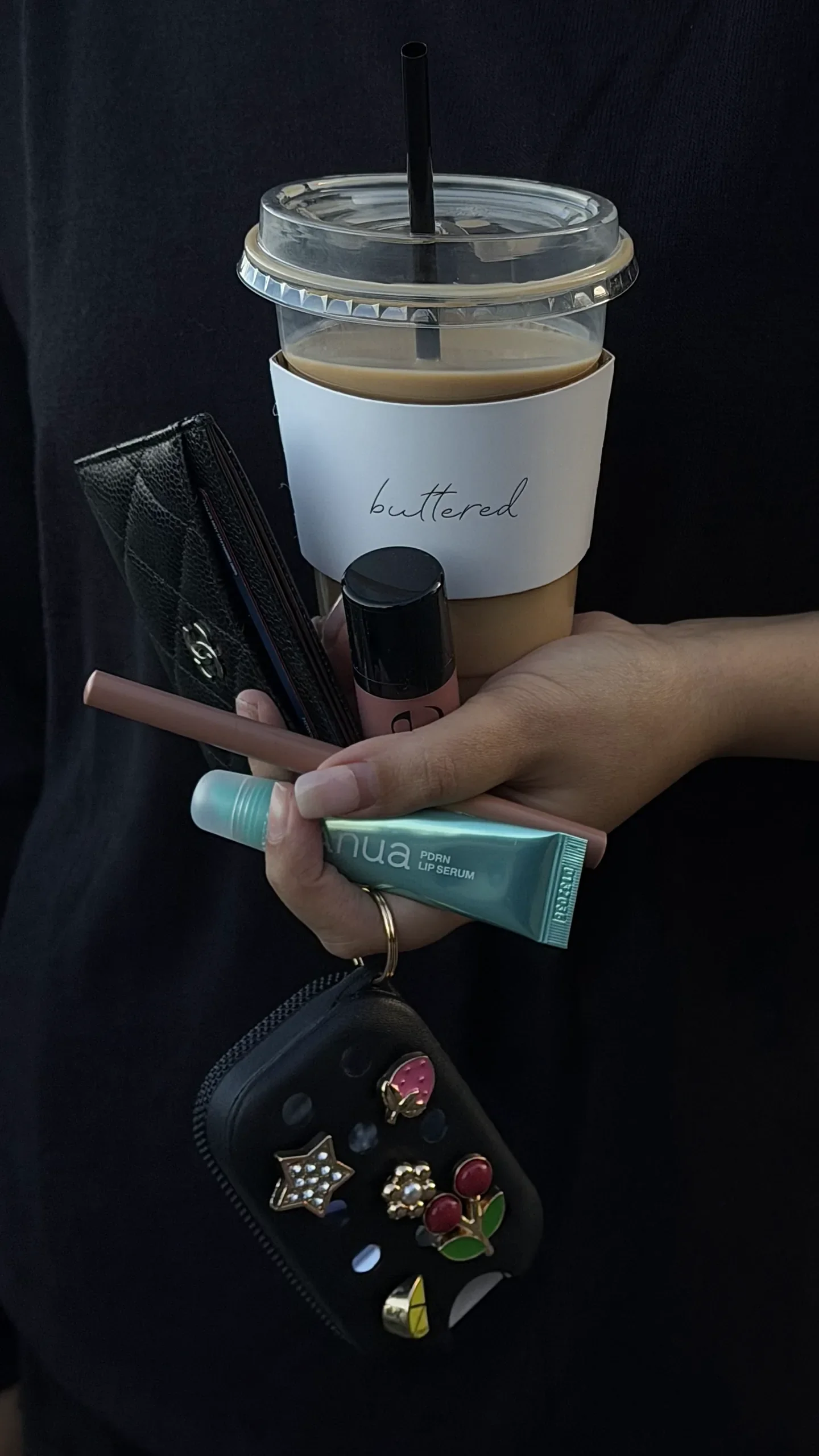 An iced coffee in a Buttered cup held alongside a card holder, lip products and a charm-covered case