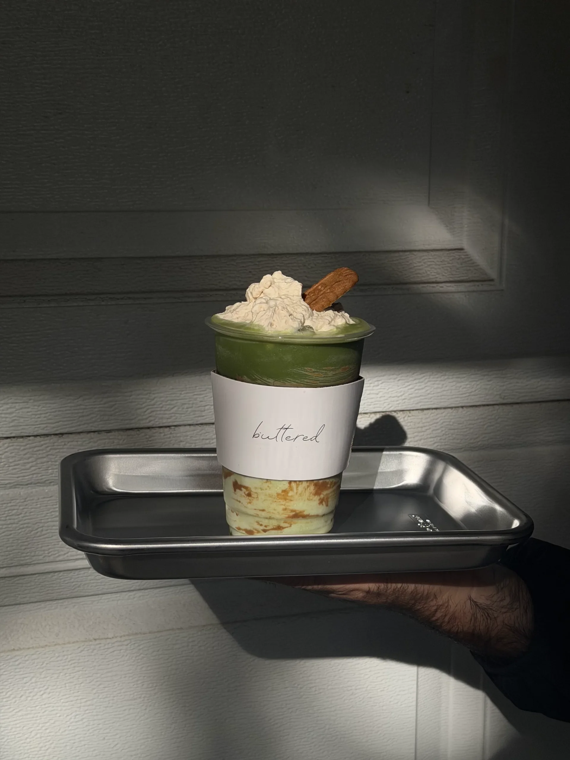 An iced matcha topped with cream and a biscuit, held out on a steel tray